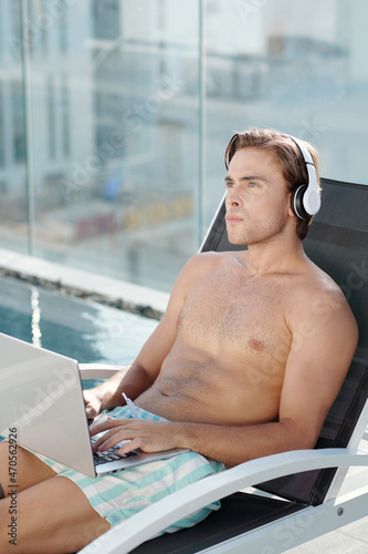 Pensive young man working on business plan when sitting on chaise-lounge by swimming pool
