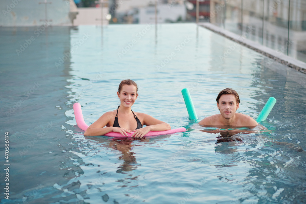Cheerful young couple swimming with pool noodles in rooftop pool
