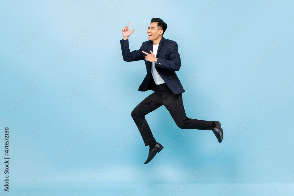 Full lenght portrait of smiling handsome Asian man jumping and pointing hands up on isolated light blue studio background