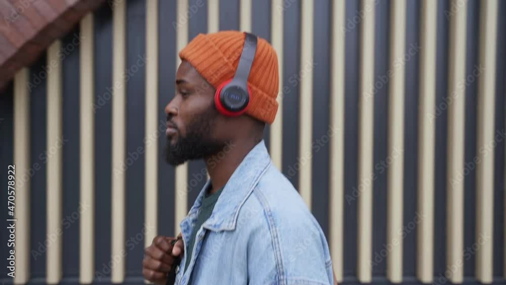 Music lover walking along street. Handsome African-American person with modern earphones walks passing large building wall in city street side view spbas