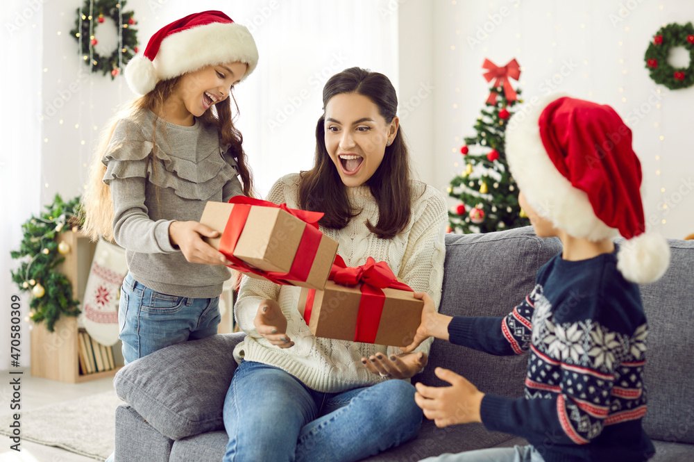 Happy family enjoying gift giving on Christmas Day. Little children prepare beautiful presents for their mother. Young woman receiving a wonderful surprise from her daughter and son in Santa caps Happy family enjoying gift giving on Christmas Day. Little children prepare beautiful presents for their mother. Young woman receiving a wonderful surprise from her daughter and son in Santa caps