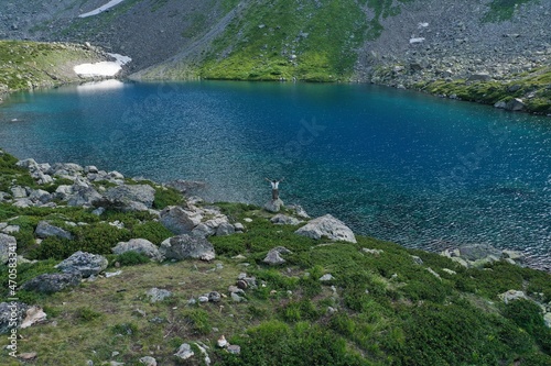 Aerial drone view of alone woman traveler in dress and hat watching on Seven colored mountain glacial lake, summer landscape: high peaks, snow, stones. Arkhyz, Caucasus, Karachay-Cherkessia, Russia