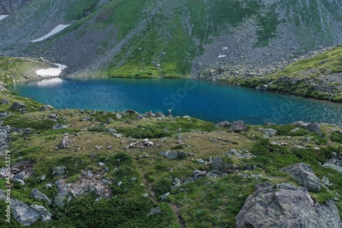 Aerial drone view of alone woman traveler in dress and hat watching on Seven colored mountain glacial lake, summer landscape: high peaks, snow, stones. Arkhyz, Caucasus, Karachay-Cherkessia, Russia