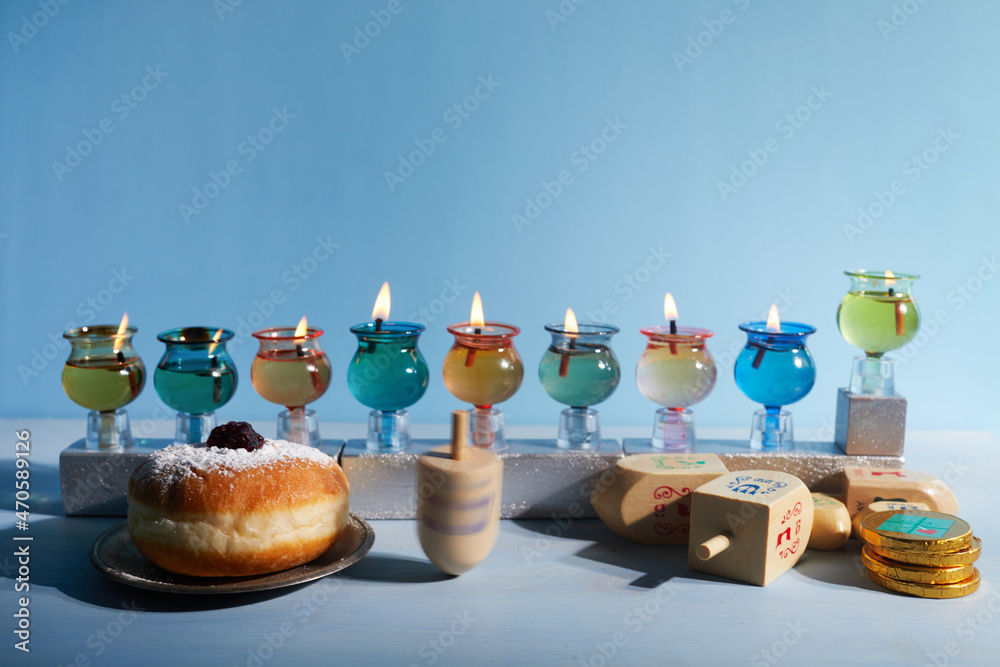 Fototapeta premium Jewish holiday Hanukkah background with oil Menorah- traditional candelabra, spinning top Dreidel and Doughnut on blue background