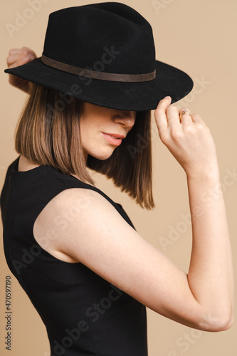Elegance portrait of a young stylish woman in a black hat. Girl posing on beige background, studio. Beautiful girl wearing classic black dress. © arthurhidden