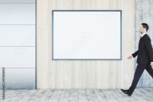 Businessman walking in abstract interior with empty wooden mock up poster on concrete tile wall and floor. Exhibition and art concept.