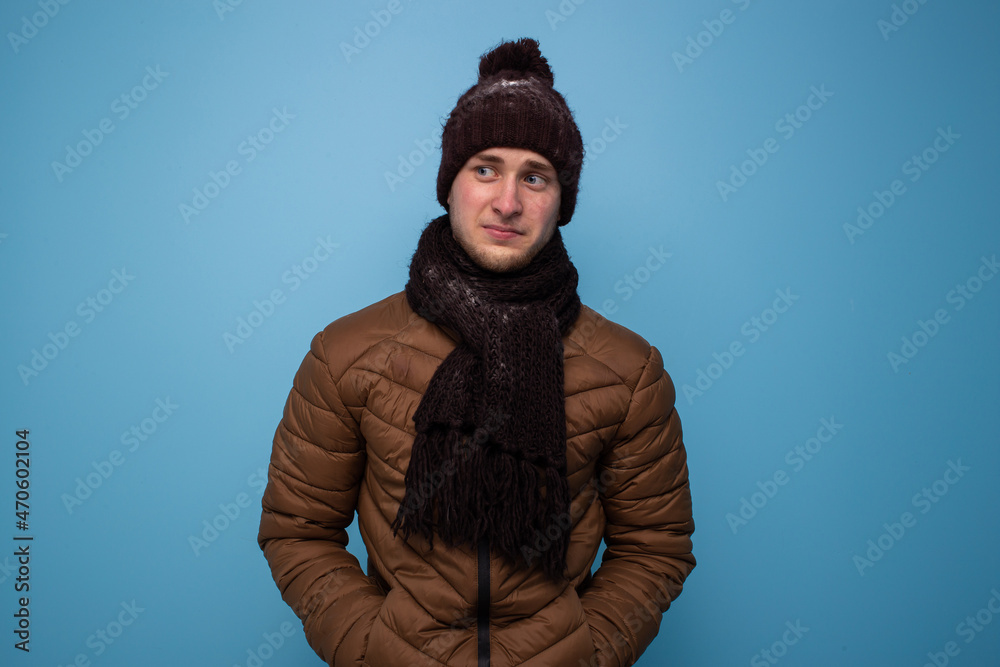 Surprised young man in brown mittens and a hat, light brown jacket on a blue background looks to the side Surprised young man in brown mittens and a hat, light brown jacket on a blue background looks to the side