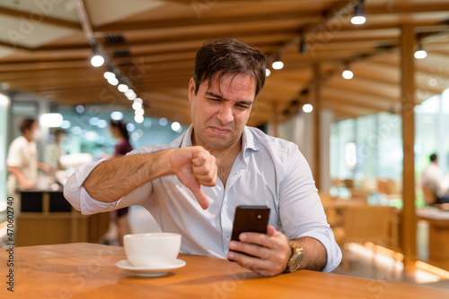 Persian man sitting at coffee shop while using mobile phone and getting bad news