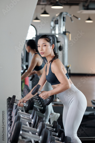 Pretty fit young Vietnamese woman choosing dummbells from stand in gym