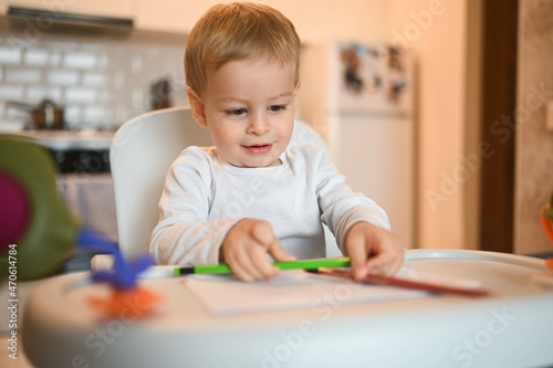 Little cute baby toddler boy blonde sitting on baby chair learning draw with colour pencils. Baby facial expressions indoors at home kitchen interior with toys. Healthy happy family childhood concept