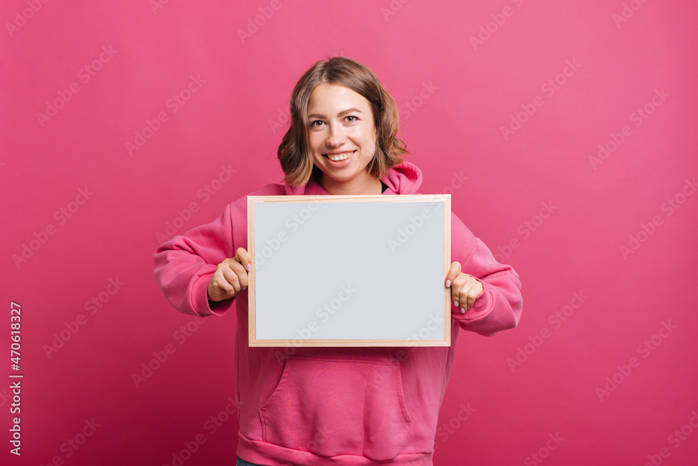 A cheerful young woman is holding a whiteboard while smiling at the camera near a pink wall A cheerful young woman is holding a whiteboard while smiling at the camera near a pink wall