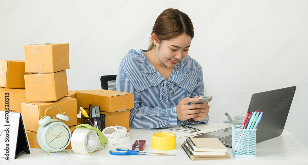 Young Asian woman business owner with many parcel boxes on the table happy online sales job, use the laptop, get an order from customers, take notes, and make arrangements for delivery by post Young Asian woman business owner with many parcel boxes on the table happy online sales job, use the laptop, get an order from customers, take notes, and make arrangements for delivery by post