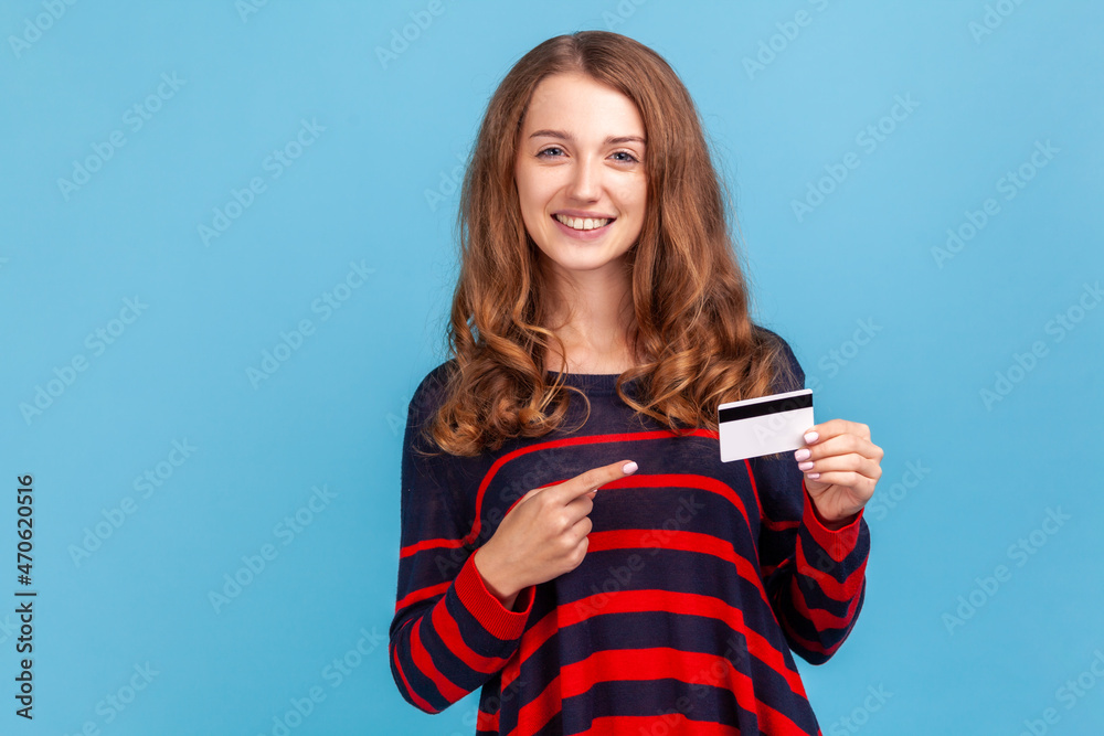 Smiling woman wearing striped casual style sweater, points finger at credit card, looking with toothy smile, recommend bank or shopping discounts. Indoor studio shot isolated on blue background. Smiling woman wearing striped casual style sweater, points finger at credit card, looking with toothy smile, recommend bank or shopping discounts. Indoor studio shot isolated on blue background.