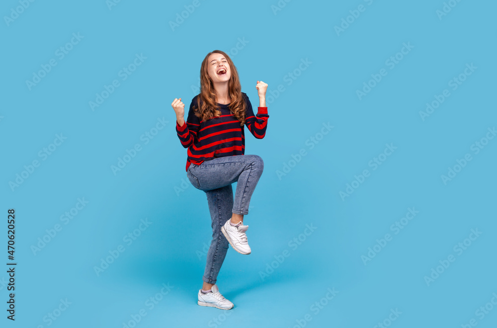 Full length positive satisfied woman wearing striped casual style sweater and jeans, raised clenched fists, being happy of winning, expressing triumph. Indoor studio shot isolated on blue background.