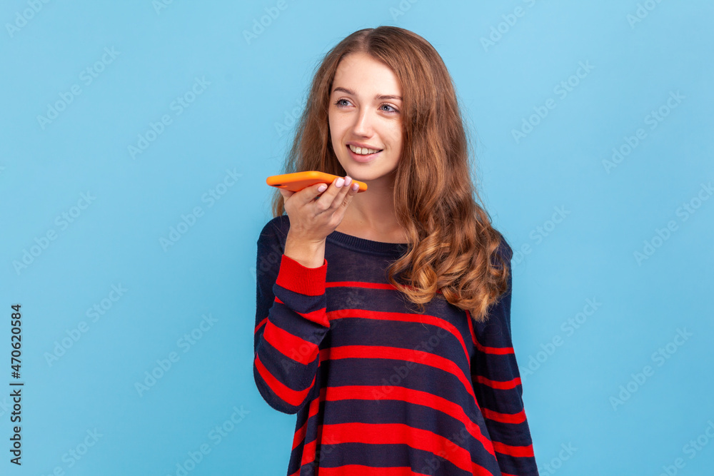 Happy young adult woman wearing striped casual style sweater, standing with smart phone, recording voice message with positive expression. Indoor studio shot isolated on blue background. Happy young adult woman wearing striped casual style sweater, standing with smart phone, recording voice message with positive expression. Indoor studio shot isolated on blue background.