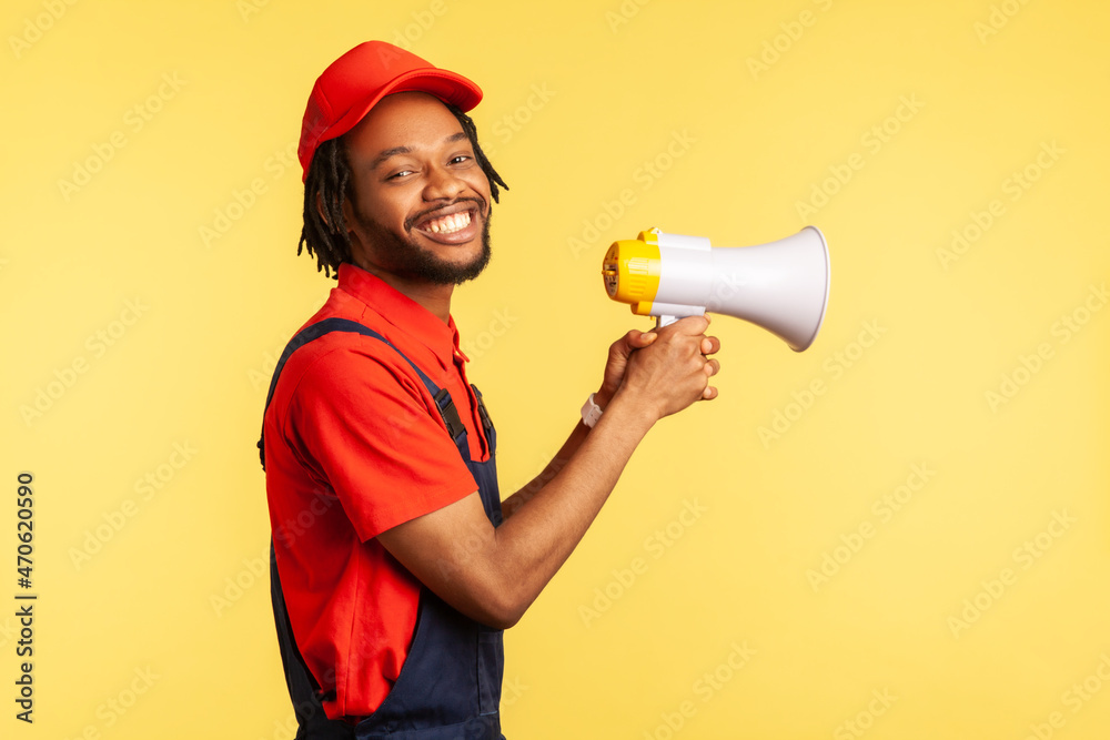 Side view portrait of smiling happy handyman wearing blue overalls and red cap, holding megaphone, looking at camera with toothy smile. Indoor studio shot isolated on yellow background. Side view portrait of smiling happy handyman wearing blue overalls and red cap, holding megaphone, looking at camera with toothy smile. Indoor studio shot isolated on yellow background.
