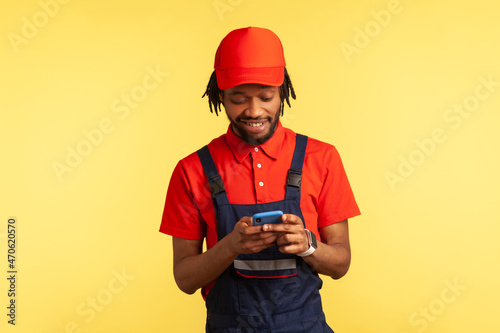 Smiling worker wearing uniform reading message on mobile phone in online delivery order app, house repair maintenance or delivery services. Indoor studio shot isolated on yellow background.