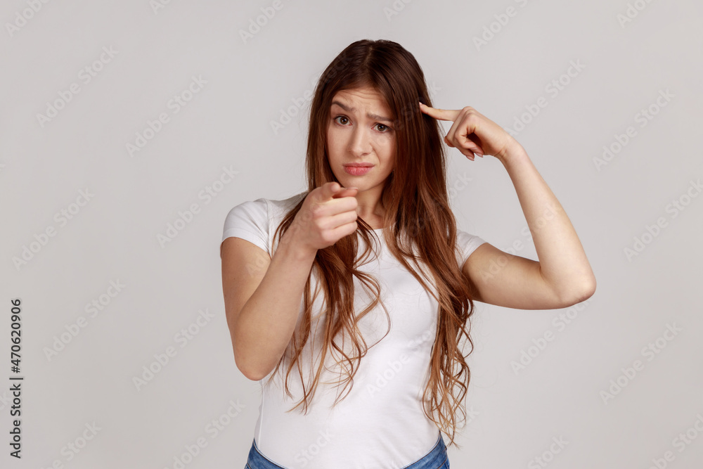 You're idiot with insane crazy plan. Displeased woman showing stupid gesture and pointing to camera, accusing of dumb idea, wearing white T-shirt. Indoor studio shot isolated on gray background. You're idiot with insane crazy plan. Displeased woman showing stupid gesture and pointing to camera, accusing of dumb idea, wearing white T-shirt. Indoor studio shot isolated on gray background.