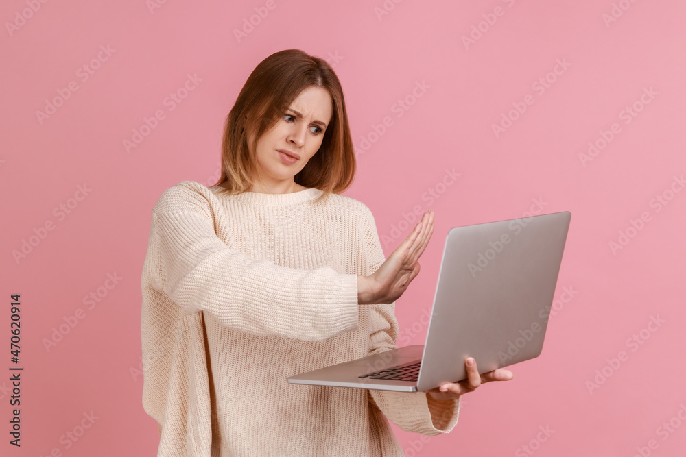 Woman showing stop gesture to laptop screen, warning with prohibition sign when talking on video call, online communication, wearing white sweater. Indoor studio shot isolated on pink background. Woman showing stop gesture to laptop screen, warning with prohibition sign when talking on video call, online communication, wearing white sweater. Indoor studio shot isolated on pink background.