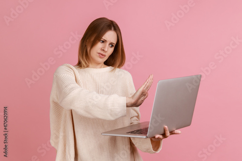 Woman showing stop gesture to laptop screen, warning with prohibition sign when talking on video call, online communication, wearing white sweater. Indoor studio shot isolated on pink background.