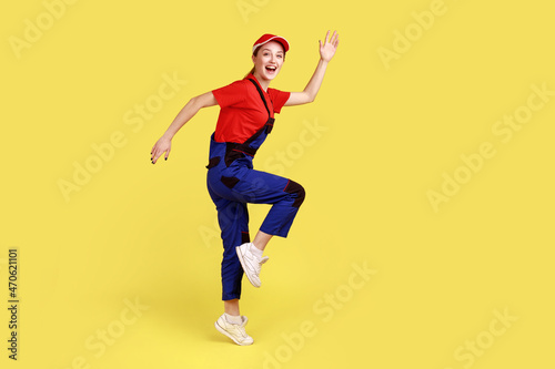 Side view portrait of excited worker woman standing on one leg with raised arms, looking at camera with happy expression, wearing overalls and red cap. Indoor studio shot isolated on yellow background © khosrork
