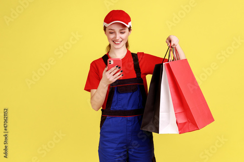 Portrait of optimistic delivery woman posing with shopping bags and smart phone in hands, using cell phone checking address, wearing overalls and cap. Indoor studio shot isolated on yellow background.
