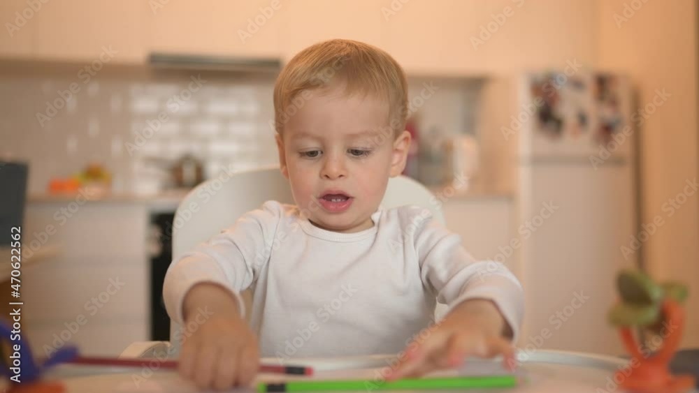 Little cute baby toddler boy blonde sitting on baby chair learning draw with colour pencils. Baby facial expressions indoors at home kitchen interior with toys. Healthy happy family childhood concept Little cute baby toddler boy blonde sitting on baby chair learning draw with colour pencils. Baby facial expressions indoors at home kitchen interior with toys. Healthy happy family childhood concept