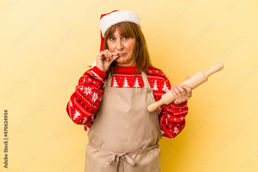 Middle age caucasian woman cooking cookies for christmas isolated on yellow background with fingers on lips keeping a secret.