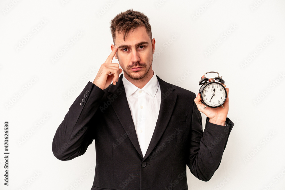 Young caucasian business man holding alarm clock isolated on white background pointing temple with finger, thinking, focused on a task. Young caucasian business man holding alarm clock isolated on white background pointing temple with finger, thinking, focused on a task.