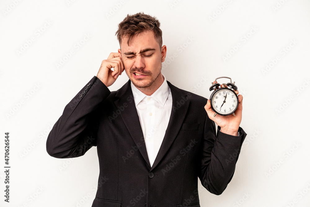 Young caucasian business man holding alarm clock isolated on white background covering ears with hands. Young caucasian business man holding alarm clock isolated on white background covering ears with hands.