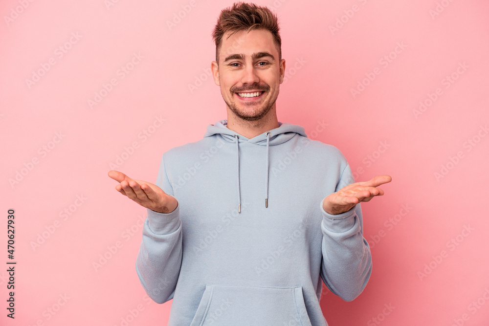 Young caucasian man isolated on pink background makes scale with arms, feels happy and confident.