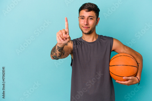 Young caucasian man playing basketball isolated on blue background  showing number one with finger. © Asier