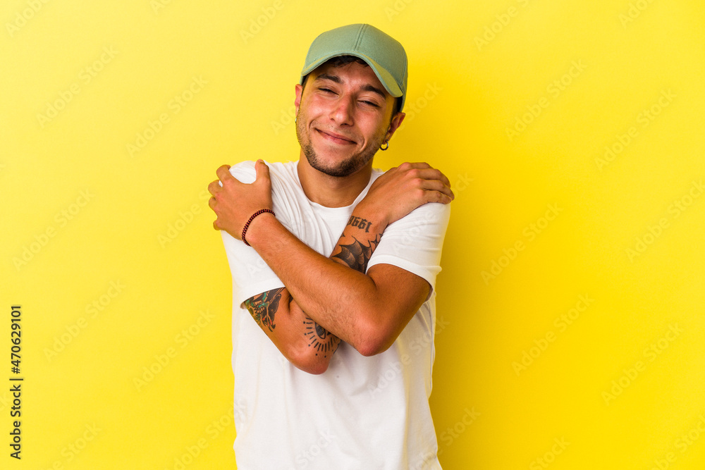 Young caucasian man with tattoos isolated on yellow background  hugs, smiling carefree and happy.