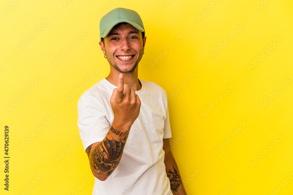 Young caucasian man with tattoos isolated on yellow background  pointing with finger at you as if inviting come closer.