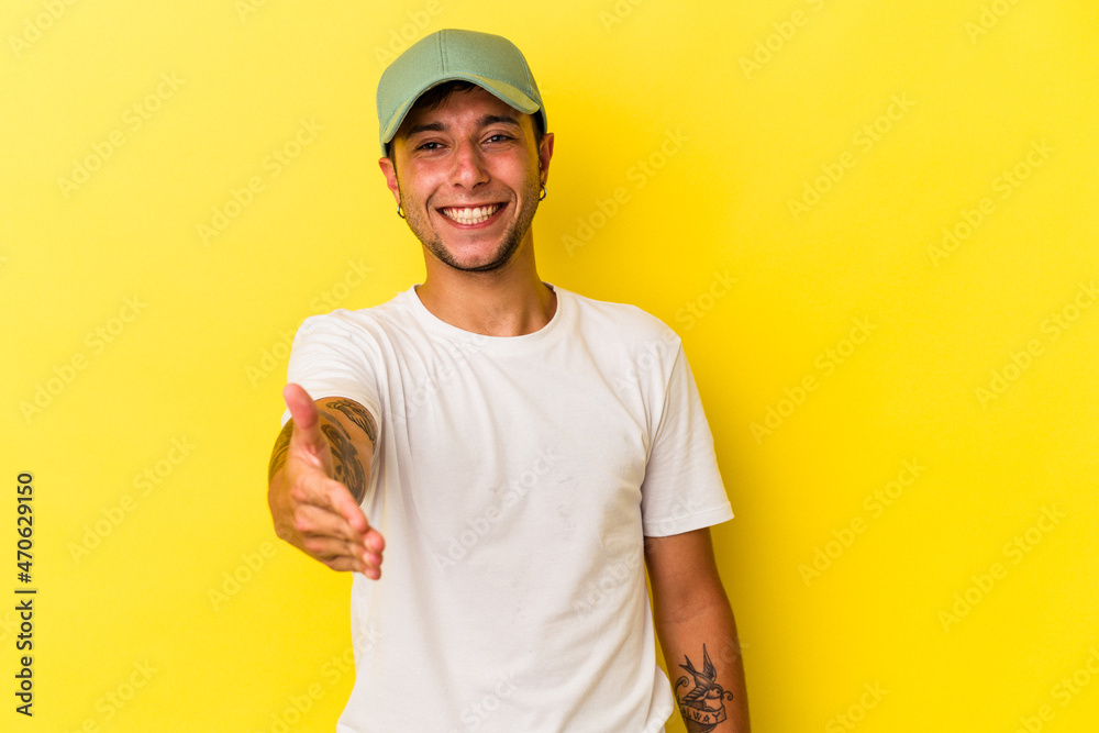 Young caucasian man with tattoos isolated on yellow background  stretching hand at camera in greeting gesture.