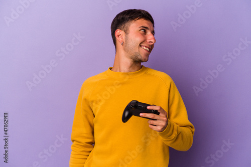 Young caucasian man holding game controller isolated on purple background looks aside smiling, cheerful and pleasant.