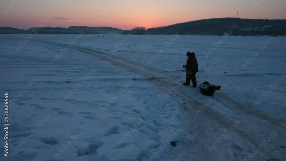 Winter fishing in Vladivostok. A fisherman walks with a fishing sled on the frozen sea at dawn. Winter fishing in Vladivostok. A fisherman walks with a fishing sled on the frozen sea at dawn.