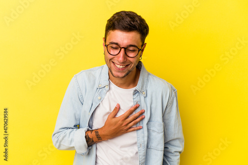 Young caucasian man with tattoos isolated on yellow background laughs out loudly keeping hand on chest.