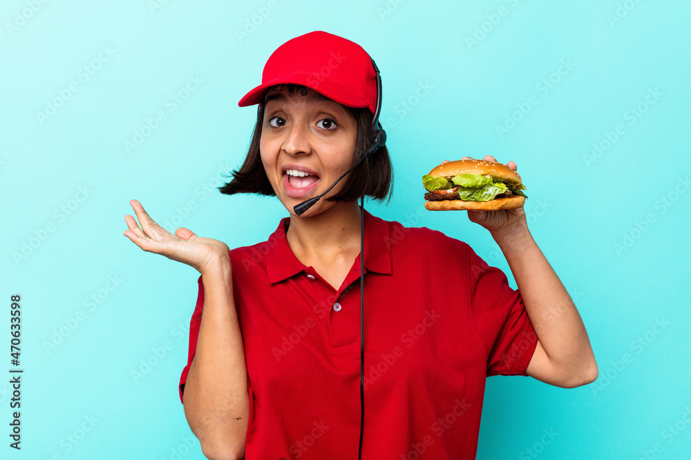 Young mixed race woman fast food restaurant worker holding a burger isolated on blue background Young mixed race woman fast food restaurant worker holding a burger isolated on blue background