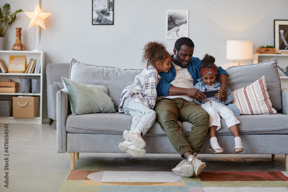Full length portrait of happy African-American father relaxing on sofa with two cute daughters and using smartphone, copy space