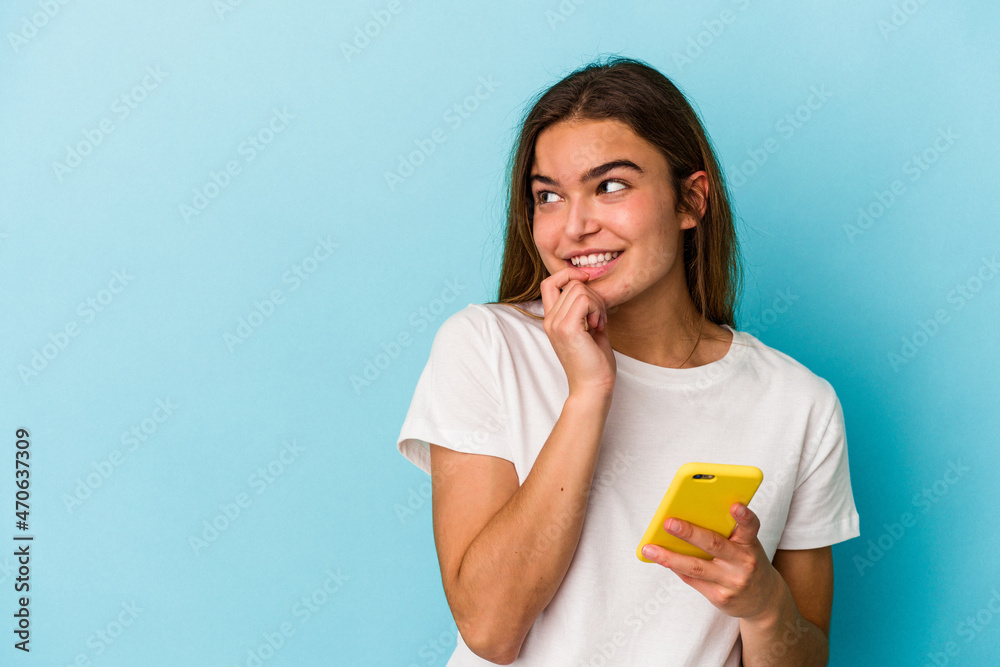 Young caucasian woman holding a mobile phone isolated on blue background relaxed thinking about something looking at a copy space.