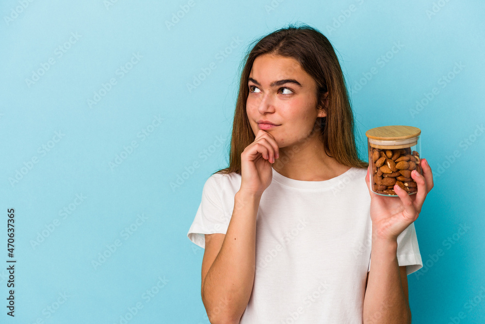 Young caucasian woman holding an almond jar isolated on blue background looking sideways with doubtful and skeptical expression. Young caucasian woman holding an almond jar isolated on blue background looking sideways with doubtful and skeptical expression.