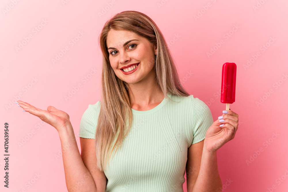 Young russian woman holding an ice cream isolated on pink background showing a copy space on a palm and holding another hand on waist. Young russian woman holding an ice cream isolated on pink background showing a copy space on a palm and holding another hand on waist.