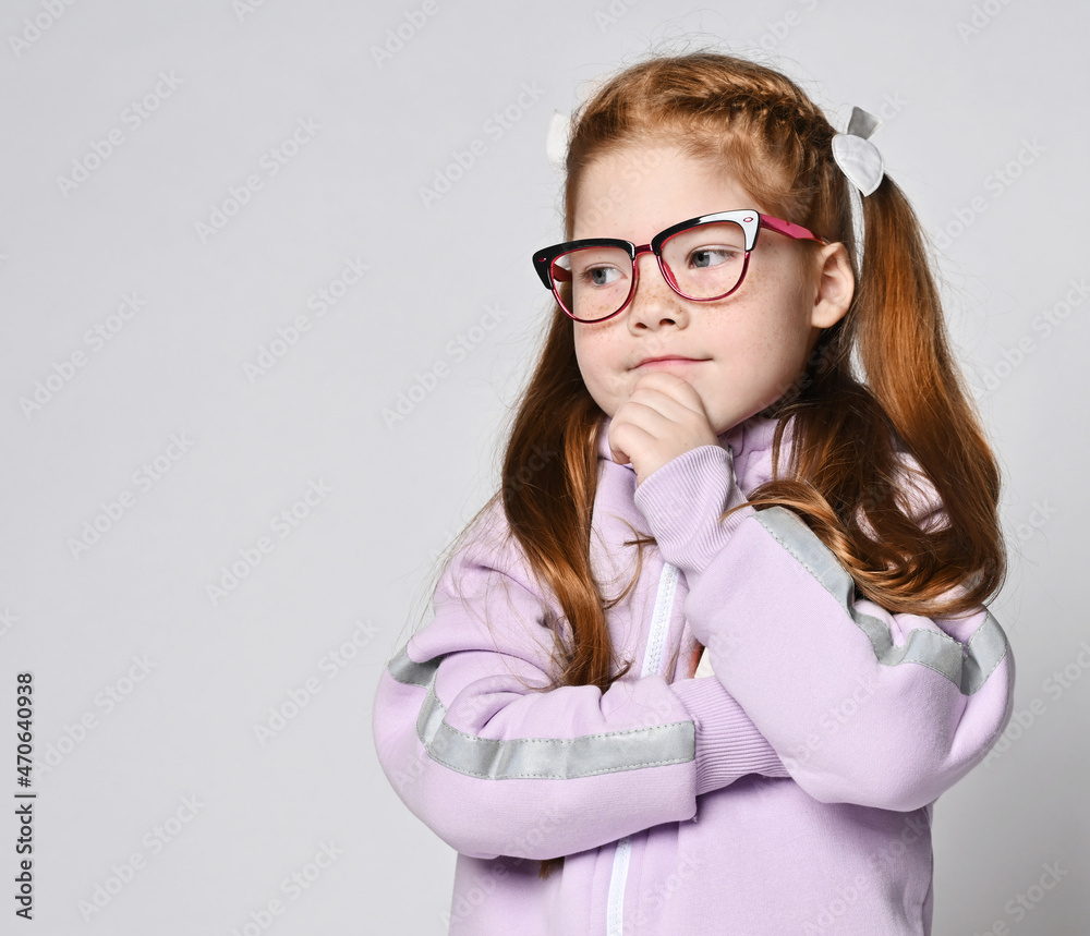 Portrait of smart redhead kid girl in glasses and pink jumpsuit with hood or hoodie with zipper standing holding hand at her chin, thinking, looking aside at copy space over light background Portrait of smart redhead kid girl in glasses and pink jumpsuit with hood or hoodie with zipper standing holding hand at her chin, thinking, looking aside at copy space over light background