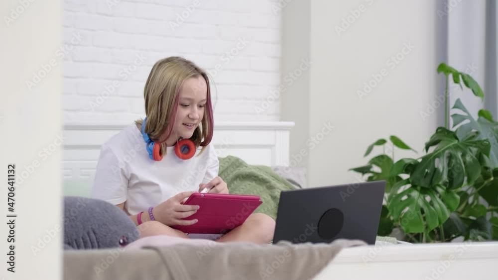 teenage girl with pink hair learns to draw from video on the internet. cute girl doing homework while sitting on the bed in the bedroom teenage girl with pink hair learns to draw from video on the internet. cute girl doing homework while sitting on the bed in the bedroom
