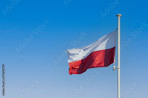 The flag of Poland flying in the wind on a tall pole against a blue sky.