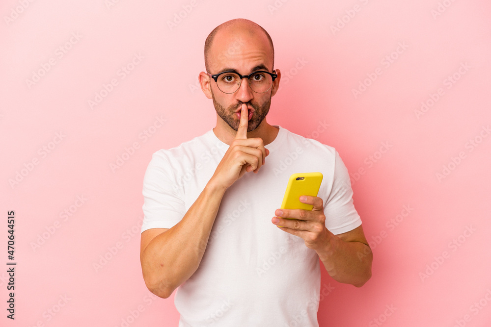 Fototapeta premium Young caucasian bald man holding mobile phone isolated on pink background keeping a secret or asking for silence.