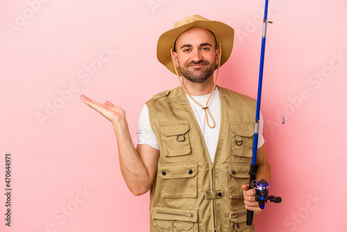 Young bald fisherman holding a rod isolated on pink background showing a copy space on a palm and holding another hand on waist.