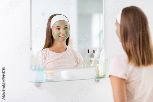 beauty, skin care and people concept - teenage girl with clay mask on her face looking in mirror at bathroom