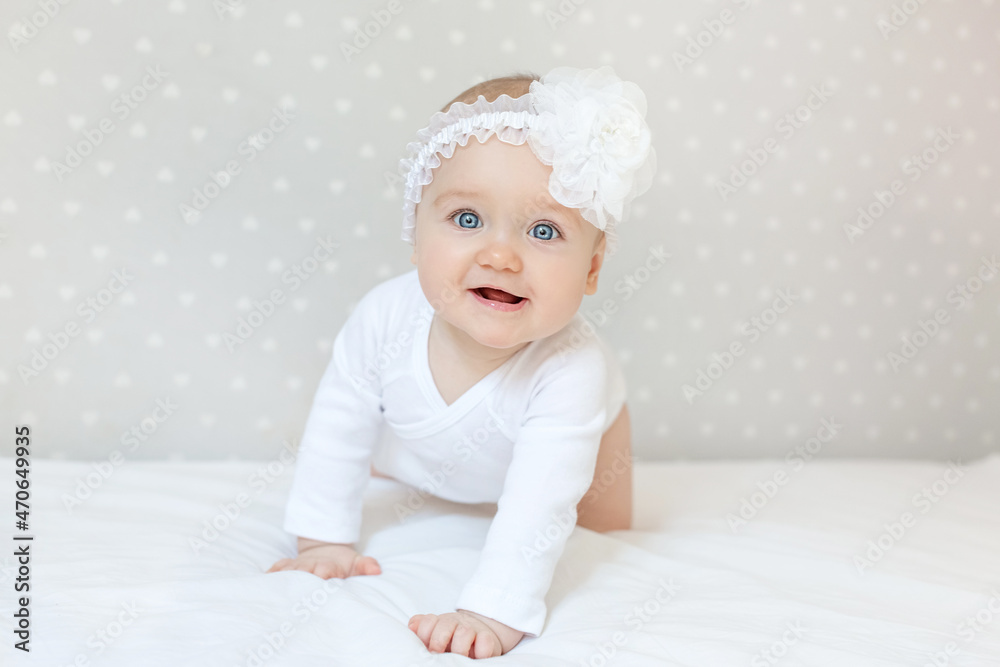 Happy toddler baby girl in a white bodysuit sitting on bed at home and looking at the camera with smile. Copy space for text Happy toddler baby girl in a white bodysuit sitting on bed at home and looking at the camera with smile. Copy space for text